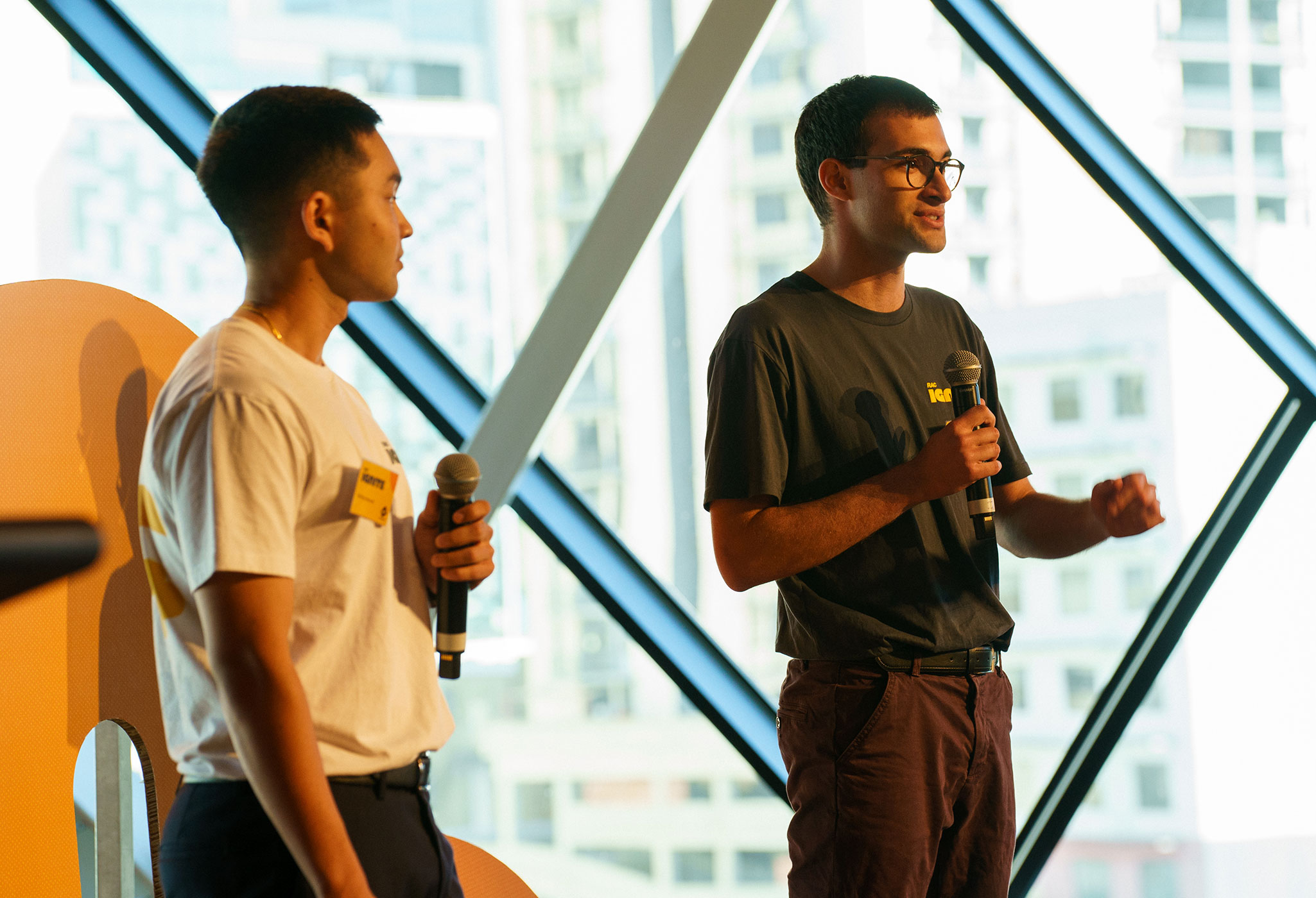 Two young men holding microphones present on stage at an RAC event, with a cityscape visible through large windows behind them.