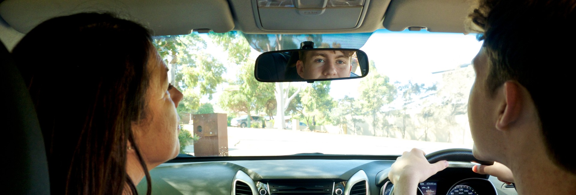A teenager is behind the wheel of a car looking in the rear vision mirror, while an adult sits in the passenger seat.