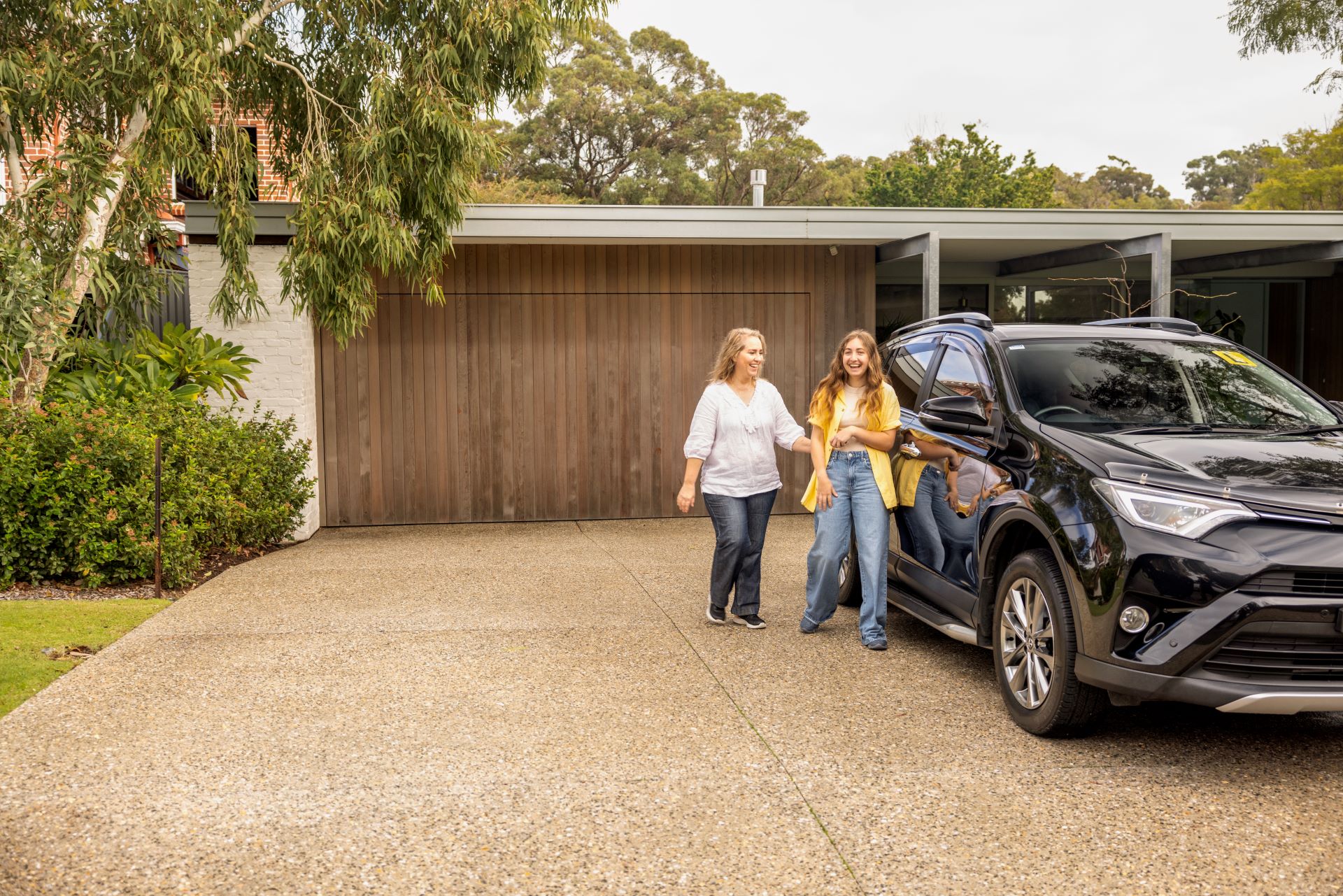 A young driver and her supervisor approach a car in their driveway. The car has yellow L plates.