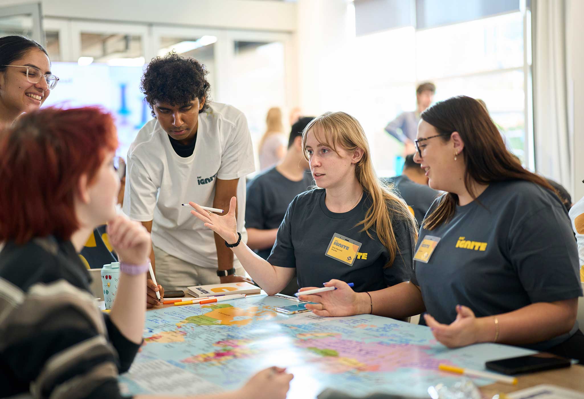 A group of young people wearing RAC Ignite branded t-shirts collaborate around a table covered with a world map during a workshop event.