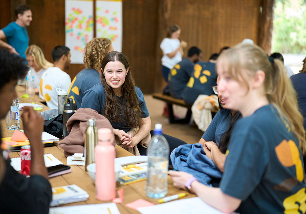 Young participants in RAC Ignite shirts sitting at wooden tables during a workshop session, smiling and chatting.