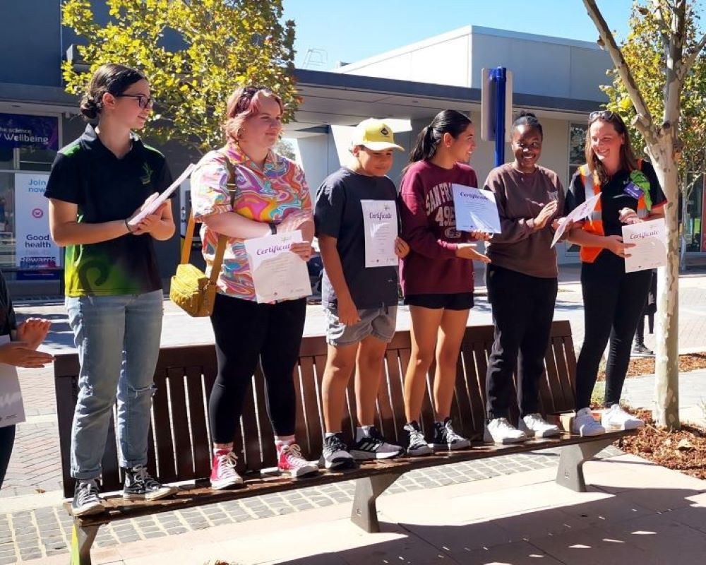Local youth stand smiling on a bench in the sunshine, showcasing their certificates, celebrating the completion of the 7 Day Makeover