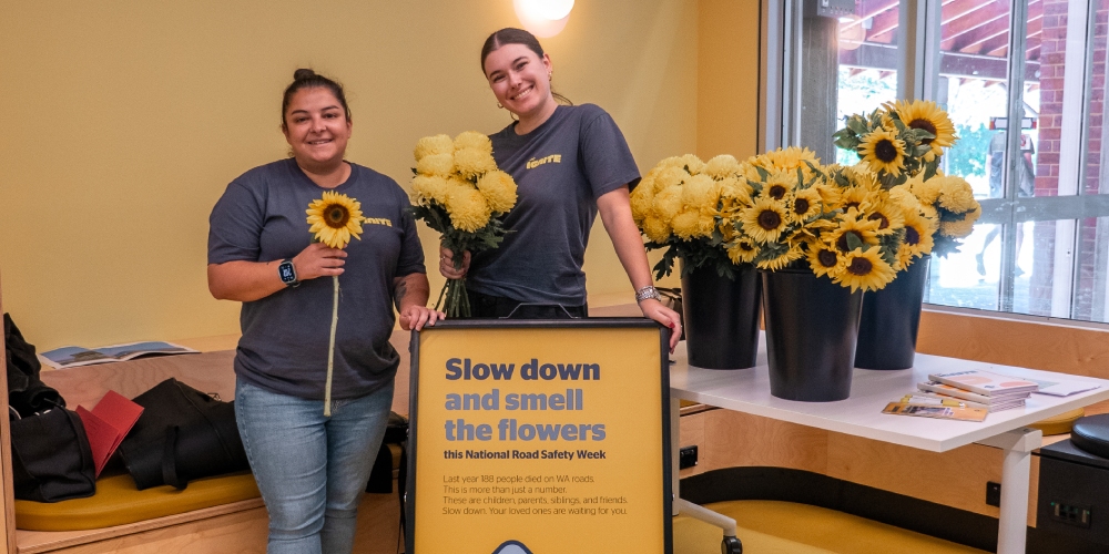 Two ignite staff at Curtin university, standing in front of a sign that reads - "slow down and smell the flowers"