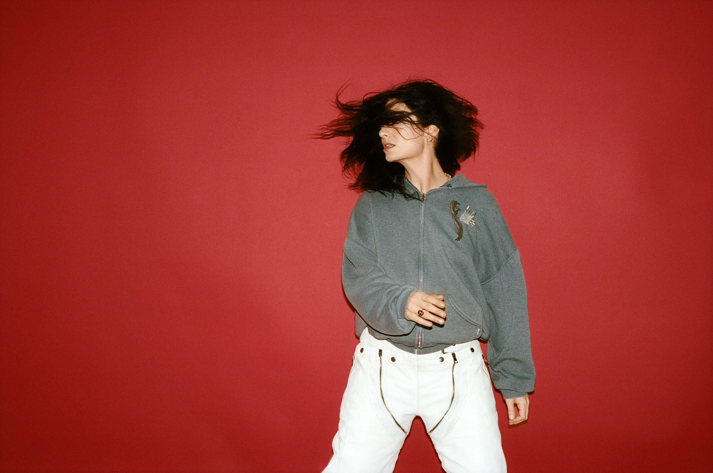 The singer Lorde is standing with a grey jumper and white pants against a bright red background.