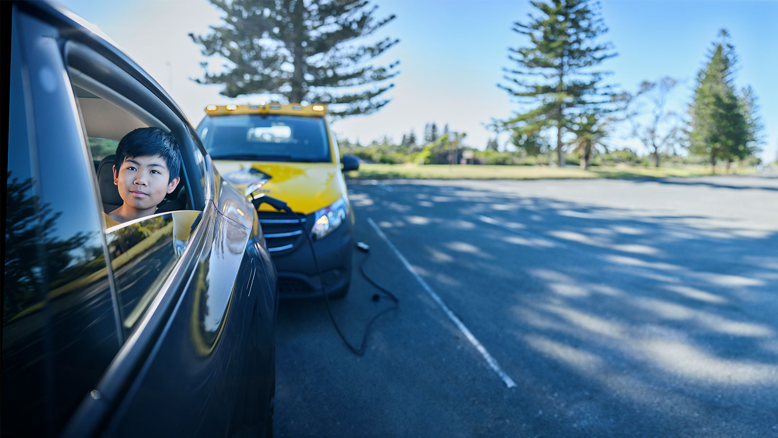 A young boy is sitting in the back seat of an electric car, looking out the open window, which an RAC patrol vehicle is charging the vehicle