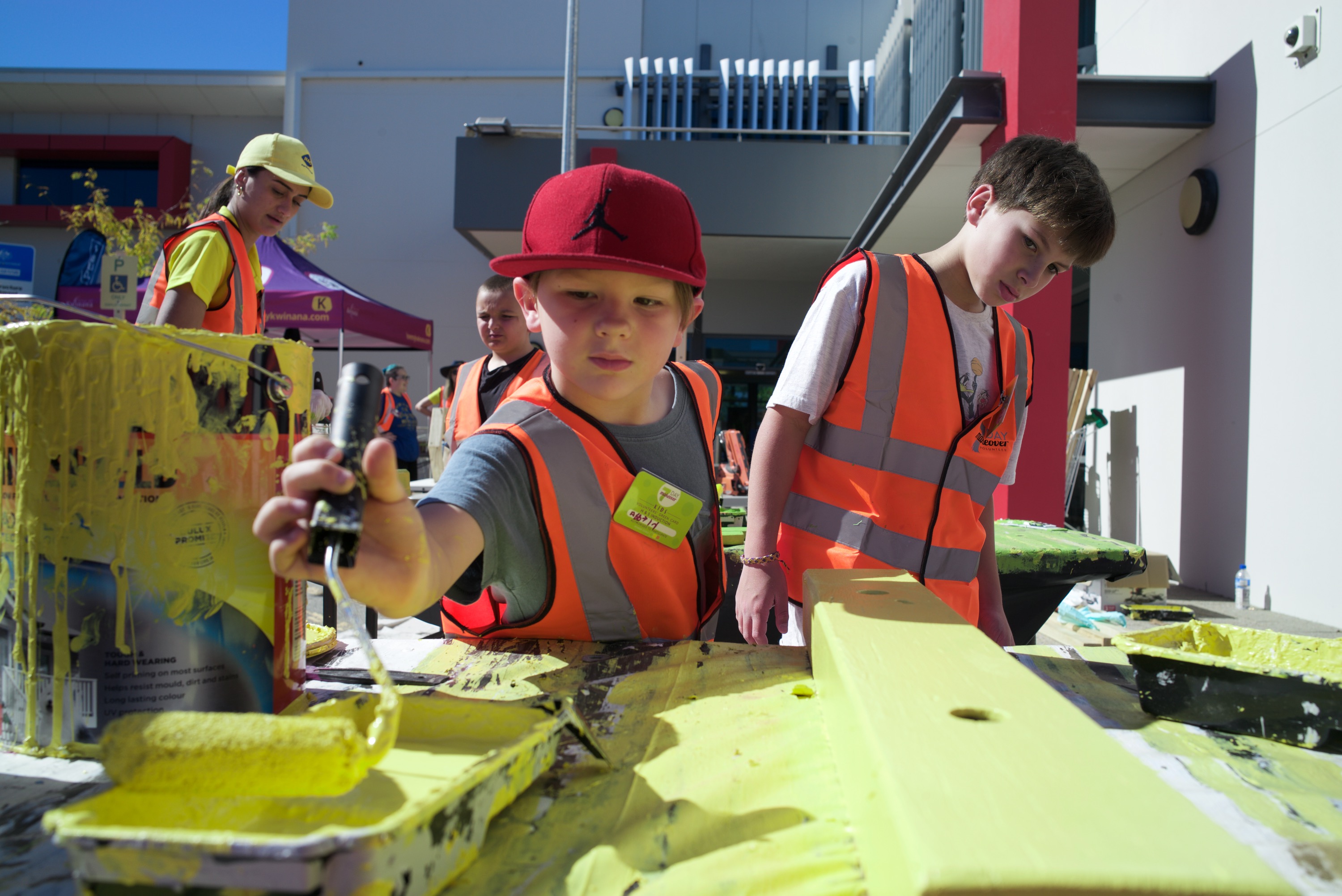 Two young boys wearing orange HIVIS jackets using bright yellow paint rollers