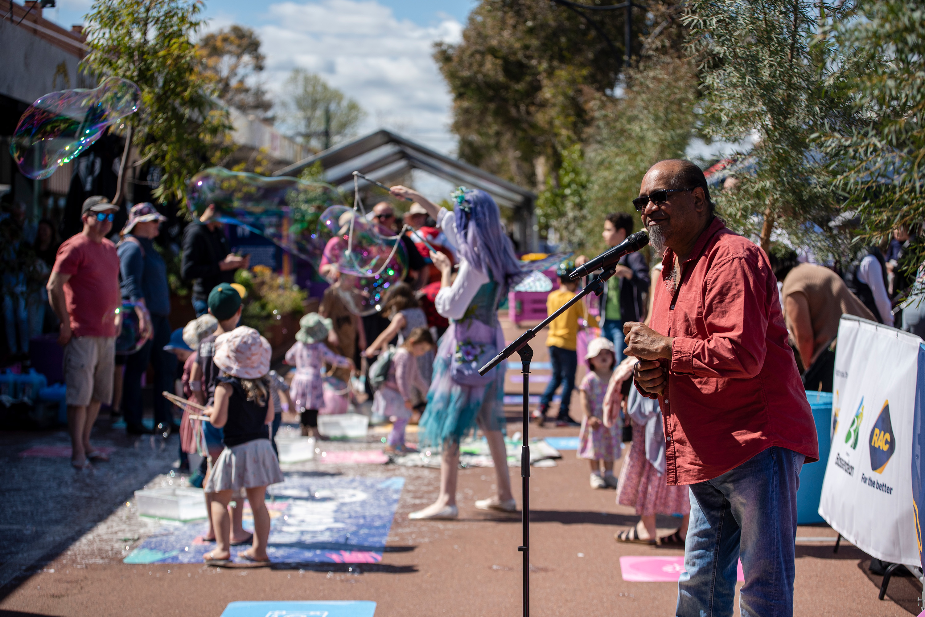 A man in a red shirt plays guitar on a street while young families gather nearby