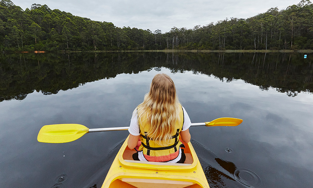 A woman is sitting in a yellow canoe, on a lake surrounded by tall trees.