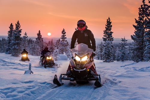 Some Aurora Village guests driving snow mobiles through deep snow with the sun setting in the background