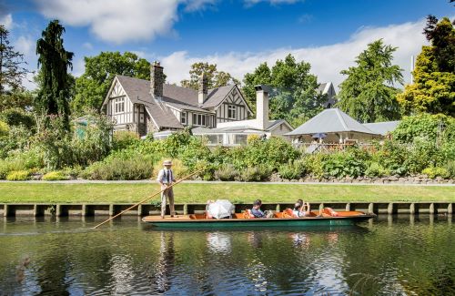 Six people sitting in a punt with one driver punting along the river in front of a heritage building surrounded by beauiful green gardens