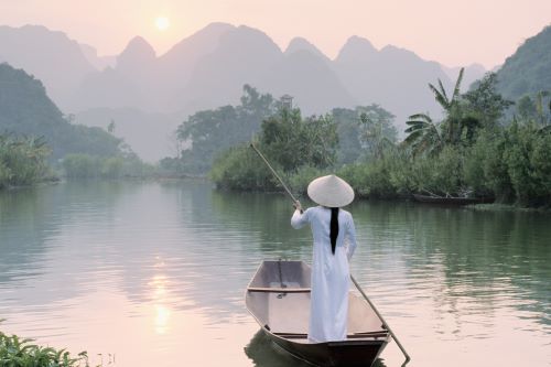 A woman in a plain white dress and typical Vietnamese hat is rowing down the river in a small wooden boat