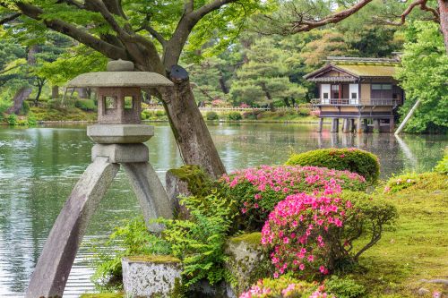 A serene Japanese garden with a stone lantern, blooming flowers, and a traditional wooden tea house by a pond