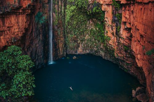 A traveller swimming in a remote gorge with a waterfall coming down the bright red rocks.
