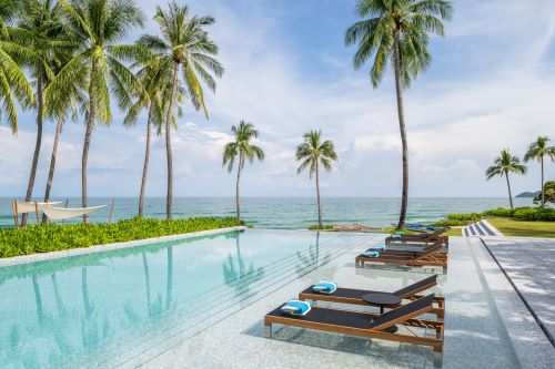Sunbeds by the pool surrounded by palm trees and the ocean in the background.