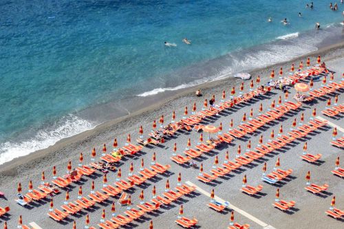 Rows of orange beach loungers and umbrellas lining a pebbled beach beside clear turquoise water.