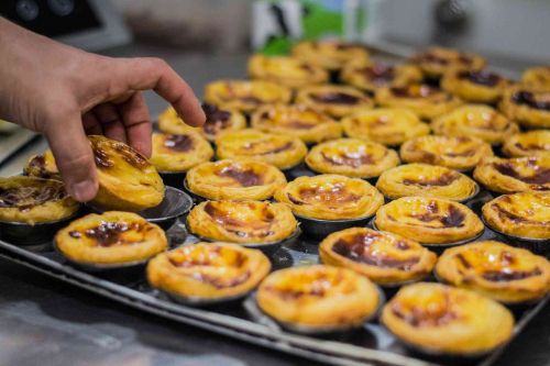 Freshly baked Portuguese custard tarts on a tray, with a hand picking up one tart.