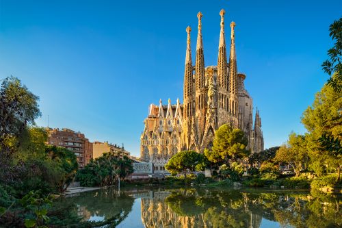 La Sagrada Família in Barcelona reflected in a pond, surrounded by trees under a clear blue sky.