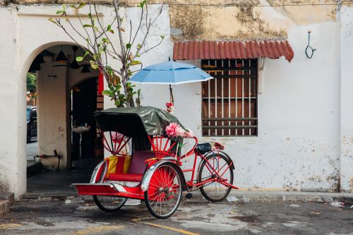 A red and black trishaw with a blue umbrella stationary against a white wall