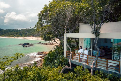 A couple of visitors standing on the balcony of their accommodation surrounded by lush nature and are looking out at the ocean