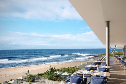 Elevated restaurant with blue-cushioned tables overlooking sandy beach and ocean, set for guests under a sunny sky.