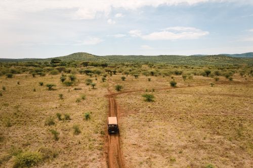 Safari vehicle driving along a dirt road through open savannah with sparse bushes and green hills in the distance.