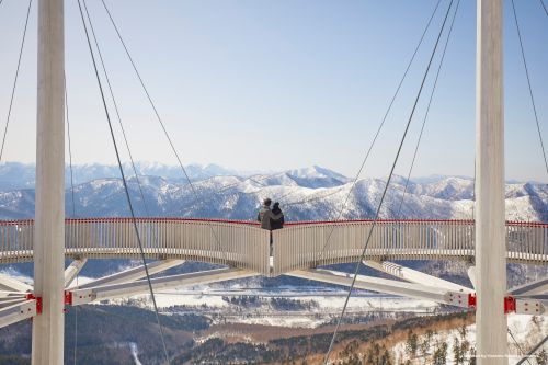 2 people standing on a suspension-style viewing platform overlooking snowy mountains