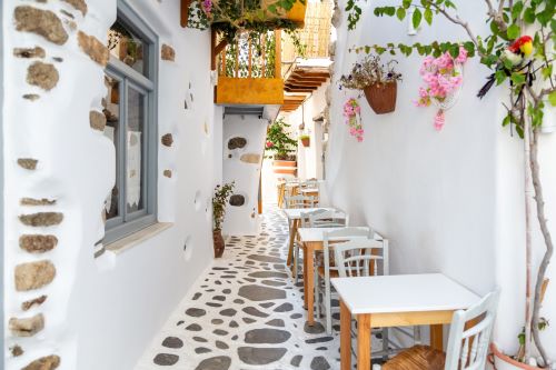 Narrow whitewashed alley with outdoor café seating and colorful flowers in a Mediterranean village.