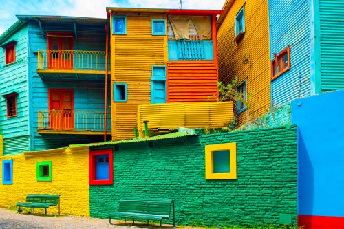 Colourful houses and buildings painted in bright shades of blue, yellow, orange and green, lining a cobblestone street with benches in the foreground.