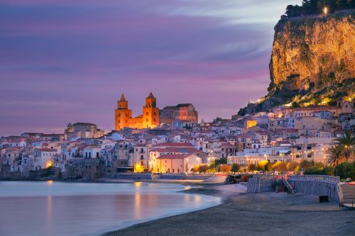Coastal view of Cefalù, Sicily, with a sandy beach, historic town, cathedral and dramatic cliff at dusk.
