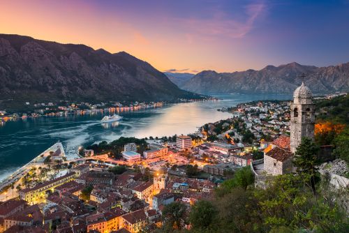 Panoramic view of Kotor, Montenegro, at dusk, with the bay, mountains, town lights and a historic stone bell tower overlooking the waterfront.