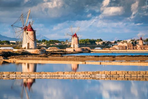 Traditional windmills and salt pans in Marsala, Sicily, reflected in still water under a cloudy sky.