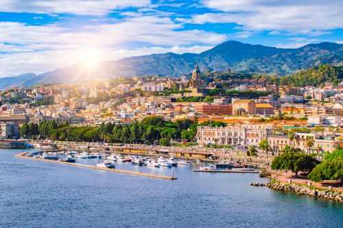 Panoramic view of Messina, Sicily, with a bustling harbour, waterfront promenade and mountains in the background.
