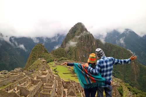 A couple stands with arms outstretched overlooking the terraced ruins of Machu Picchu, surrounded by steep green mountains and low clouds.