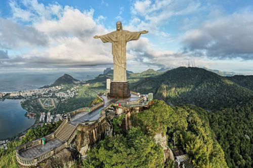 Christ the Redeemer statue stands on a mountaintop above Rio de Janeiro, with the city, coastline and forested hills visible below.