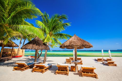 White sandy beach with wooden lounge chairs under thatched umbrellas, palm trees, and turquoise water