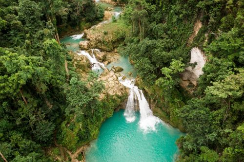 Aerial view of cascading waterfalls flowing into turquoise pools surrounded by dense green tropical forest and rocky terrain