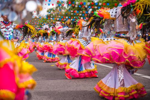 Colorful street parade with dancers in vibrant costumes featuring pink, yellow, and feathered headpieces.