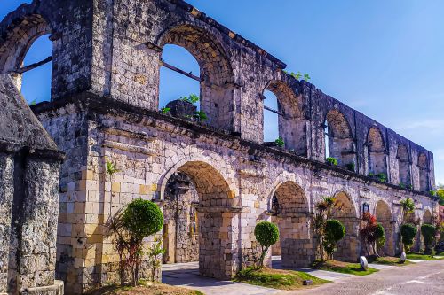 Historic stone ruins with large arched windows and doorways, partially weathered and covered with patches of greenery, under a bright blue sky with small manicured shrubs line the walkway in front of the structure
