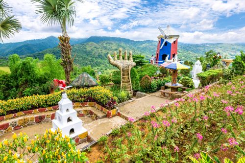 Colourful hillside garden with vibrant flowers, decorative sculptures, and a small windmill structure, overlooking lush green mountains under a partly cloudy sky