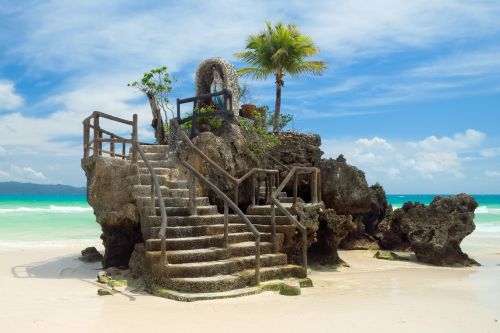Rock formation with a staircase and a small shrine, surrounded by clear blue ocean and white sand