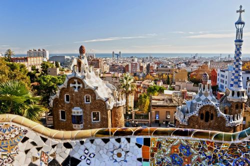 The vibrant cityscape of Barcelona, Spain, featuring the colourful and ornate mosaic tiles of Park Güell in the foreground, with a view of the city's buildings stretching out to the horizon.