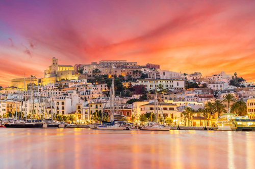 A stunning sunset over Ibiza's marina, with vibrant, colourful skies illuminating the white buildings and yachts docked along the waterfront.