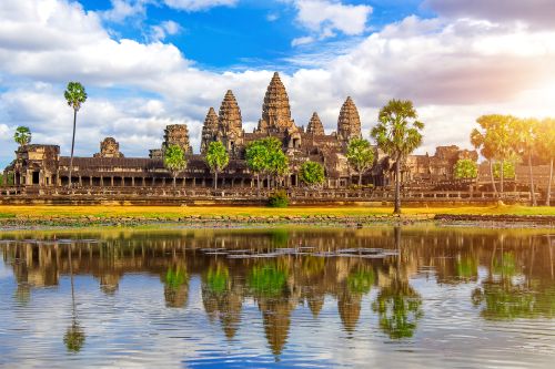 Angkor Wat temple complex reflected in a still water pool, with palm trees and warm sunlight casting a glow over the historic stone structures.