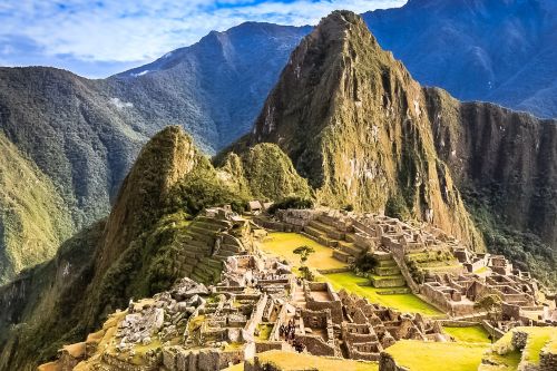 Aerial view of Machu Picchu, showing the stone ruins set among green terraces with steep mountain peaks rising in the background.