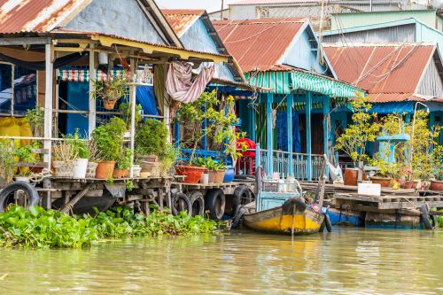 Riverside houses on stilts in Chau Doc, with colourful facades, potted plants and a small boat moored along a calm, muddy waterway.