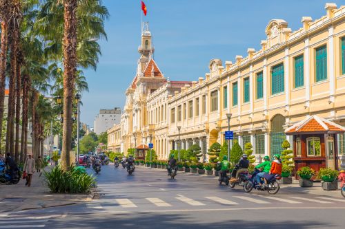 Street scene in Ho Chi Minh City with rows of historic buildings, palm trees and motorbikes travelling along a wide road under a clear blue sky.