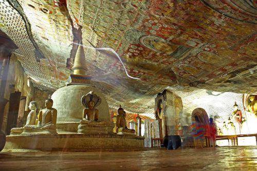 Interior of a cave temple featuring seated Buddha statues and a central stupa, with richly detailed painted ceilings and walls in warm tones, and soft light filtering across the stone floor.
