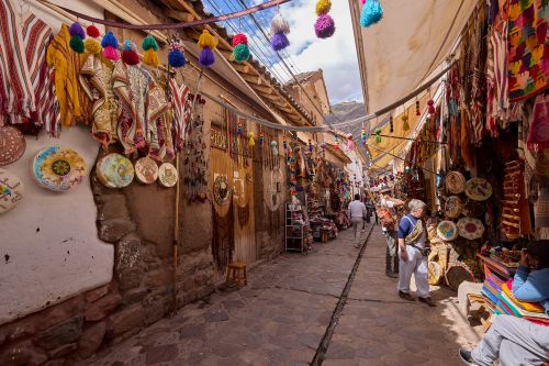 Narrow market street lined with colourful textiles, woven goods and hanging decorations, with people browsing stalls along a stone pathway in a Peruvian village setting.