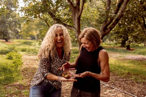 Two women surrounded by nature are laughing in the camera and holding some chestnuts