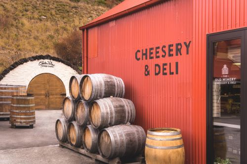 A rustic red building labelled "Cheesery & Deli" with wine barrels stacked outside, located next to a winery with a stone archway leading to a wine cellar.